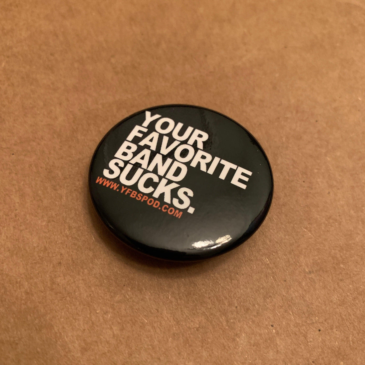 Your Favorite Band Sucks Pin, single 1.5 inch black button pin with white lettering on a wood surface