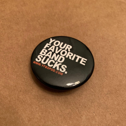 Your Favorite Band Sucks Pin, single 1.5 inch black button pin with white lettering on a wood surface