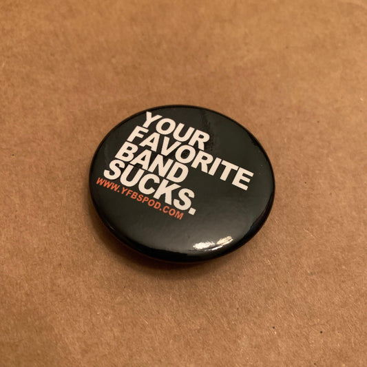 Your Favorite Band Sucks Pin, single 1.5 inch black button pin with white lettering on a wood surface